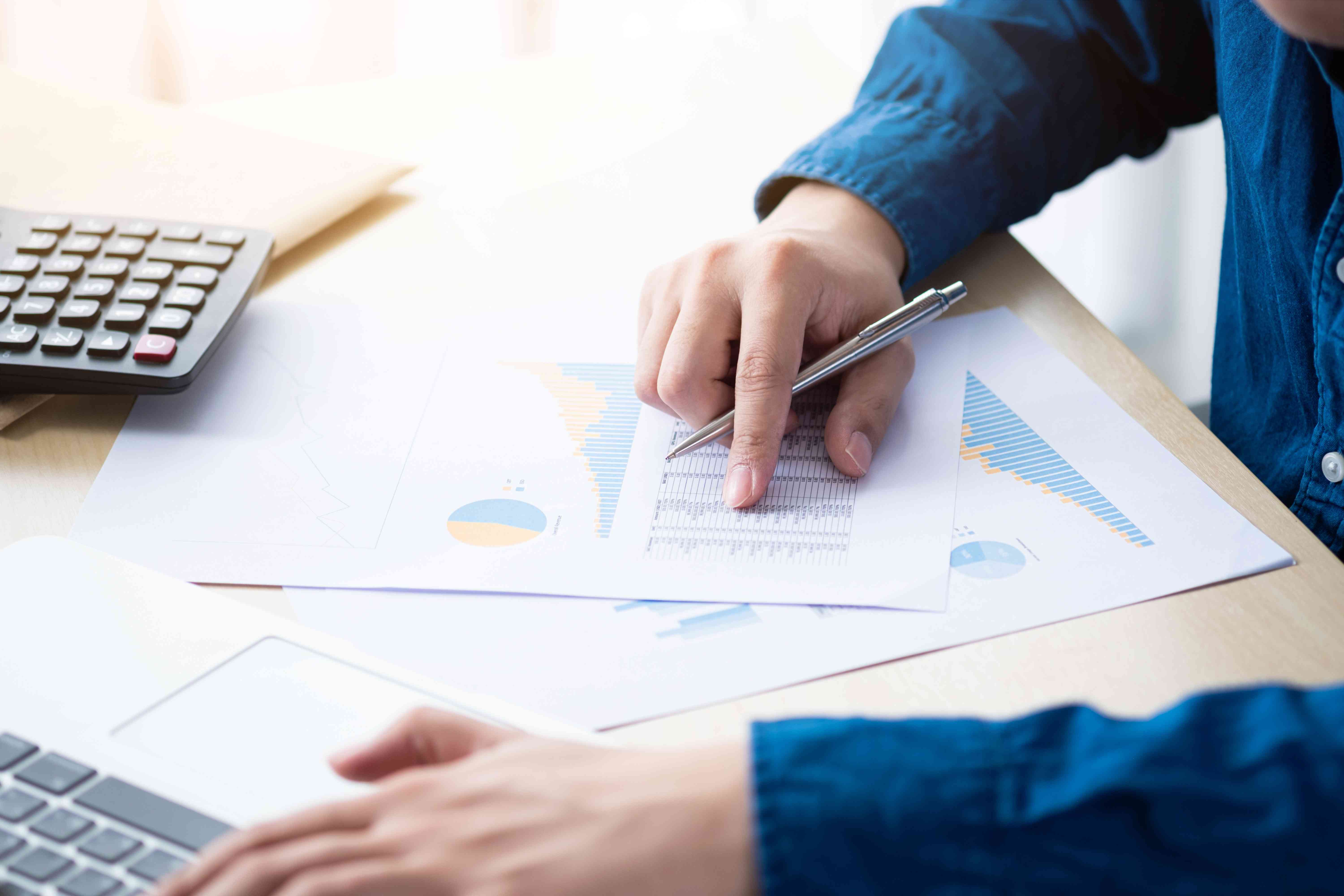 A man types on a laptop with one hand while holding a pen reviewing documents with charts in the other hand.