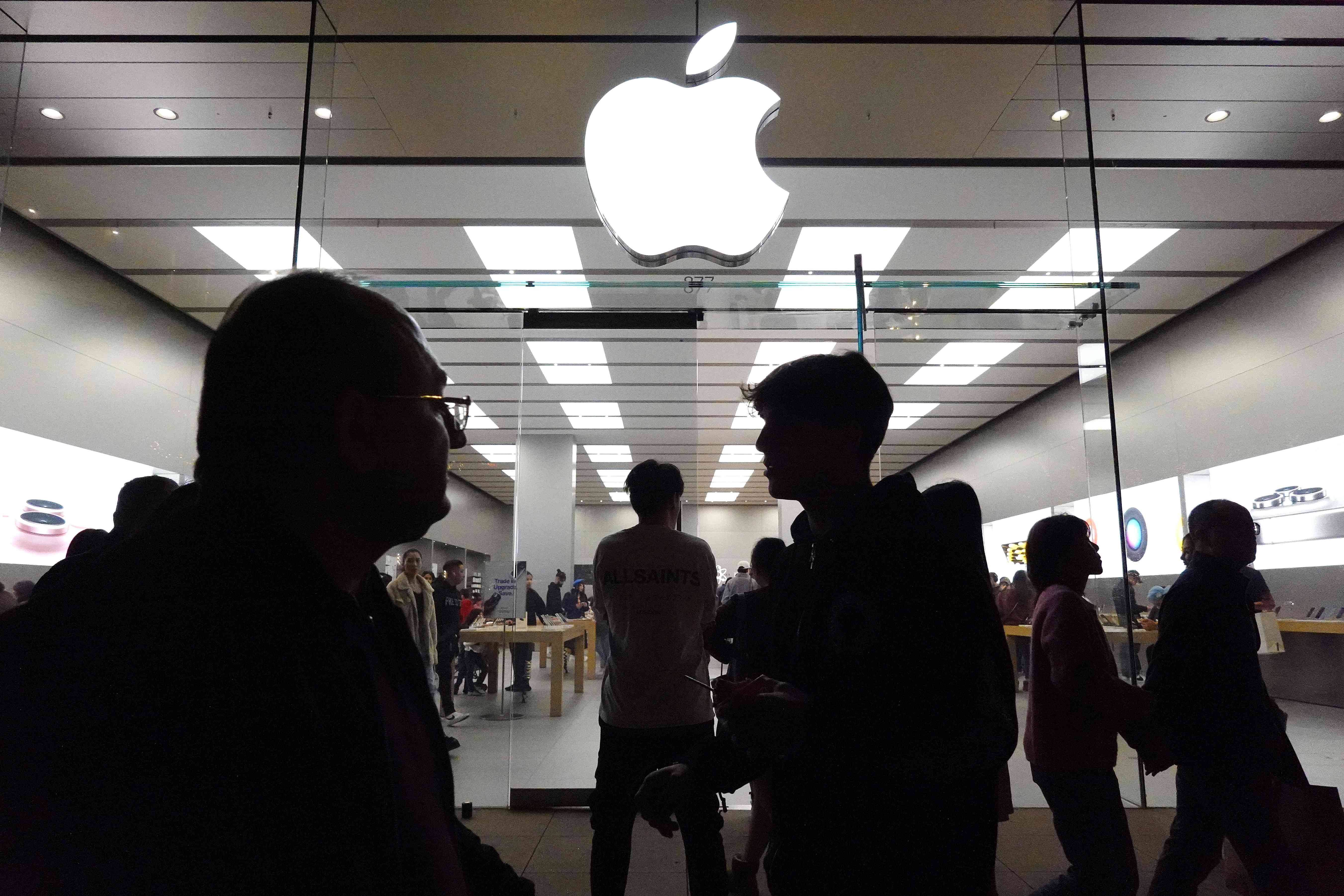 People pictured in shadow walk past an Apple store displaying an Apple logo in a shopping mall. 