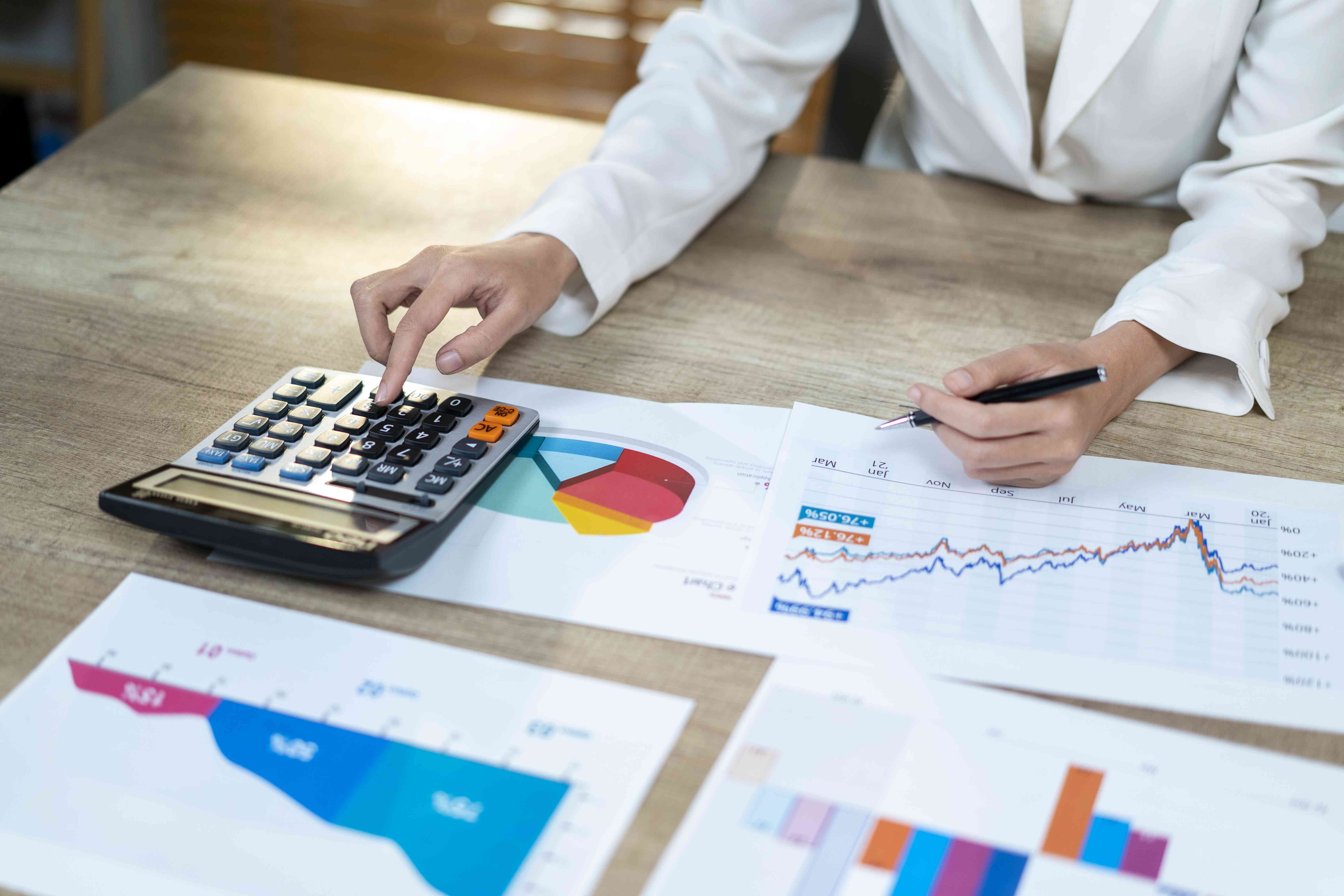 A close-up of a woman's hands using a calculator, while she sits at a desk with several documents displaying financial charts. 