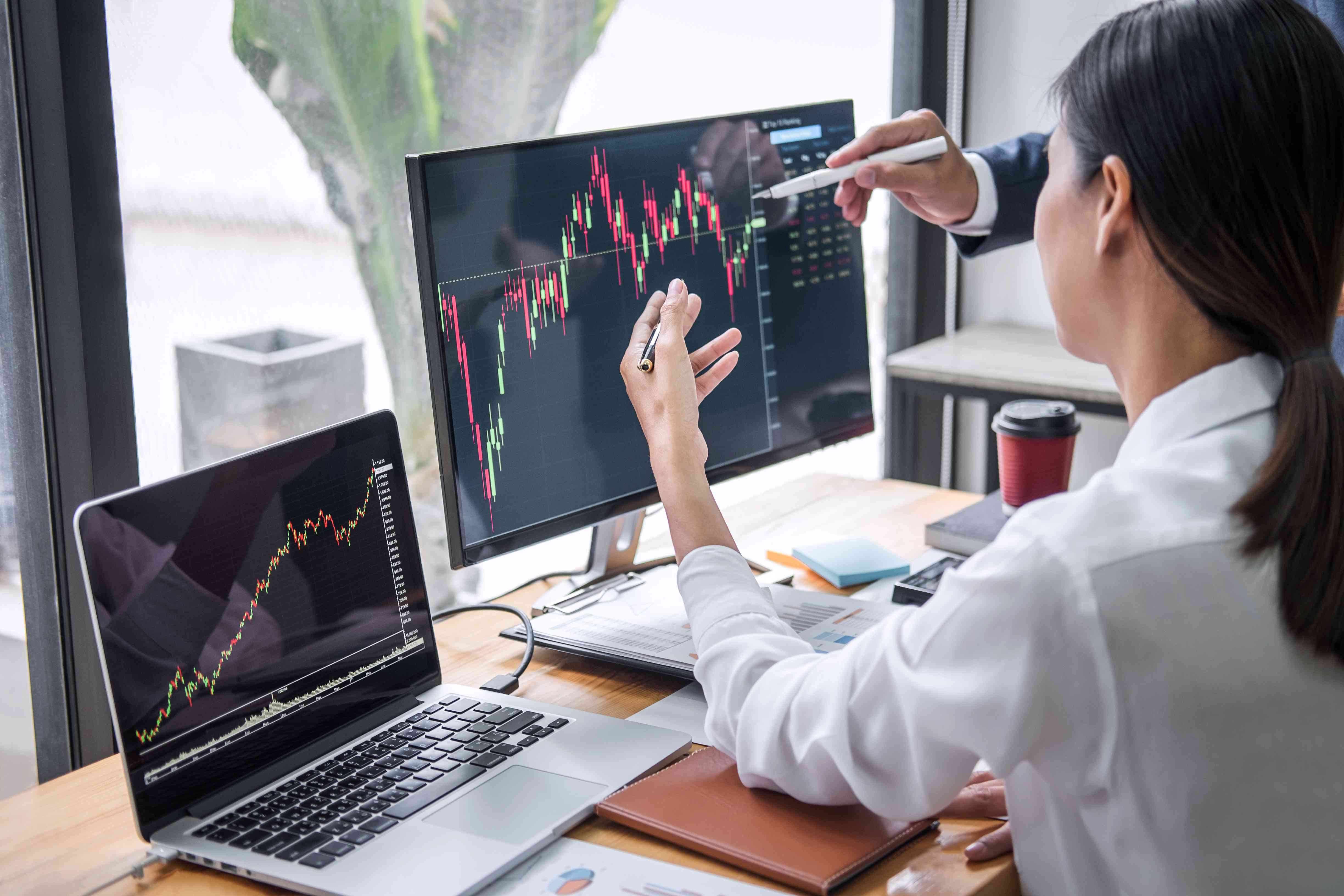 Rear View Of Businesswoman With Colleague Discussing Graph On Desktop Pc At Office