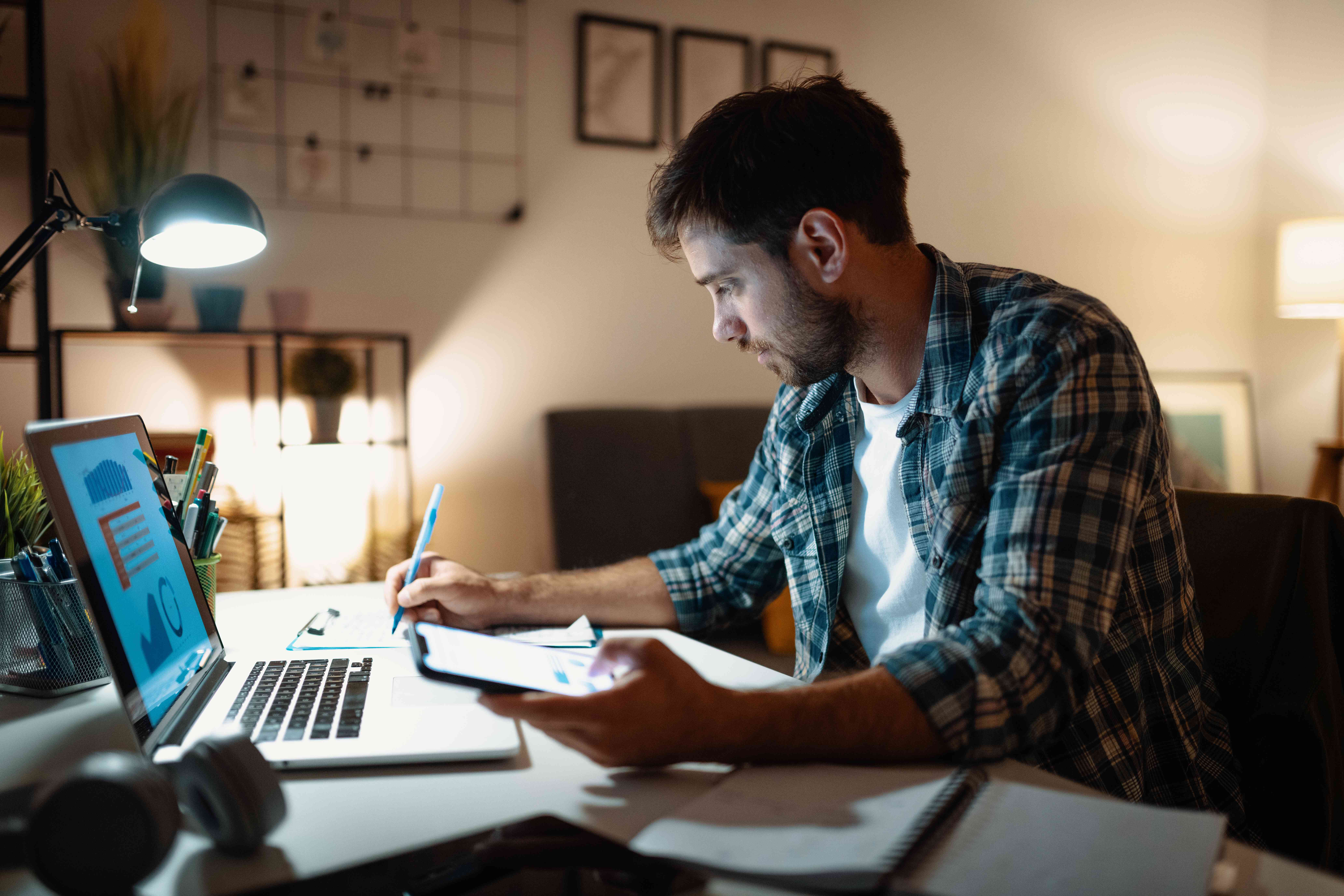 An investor works at night using a laptop and writes notes in a notebook to develop a trading strategy for the next day.